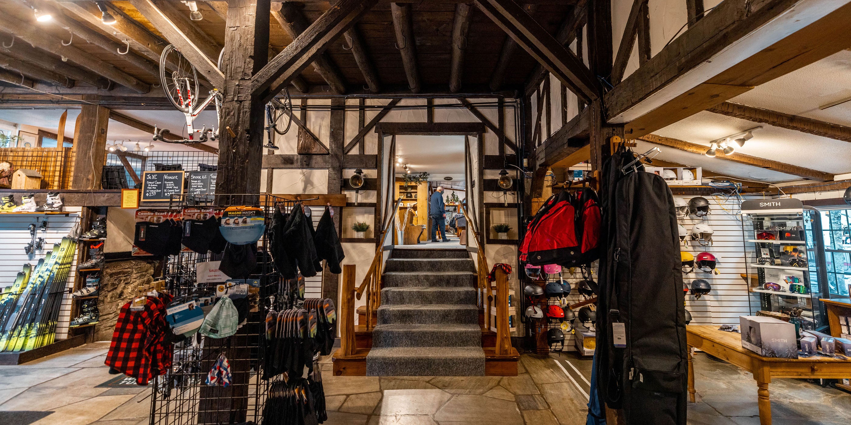 Floor of Kenver ski shop with two vertical wooden beams in the front, and a small stair case in the middle. In the foreground is a display of cold-weather masks. In the left background are skis, while in the right background are ski goggles and helmets.