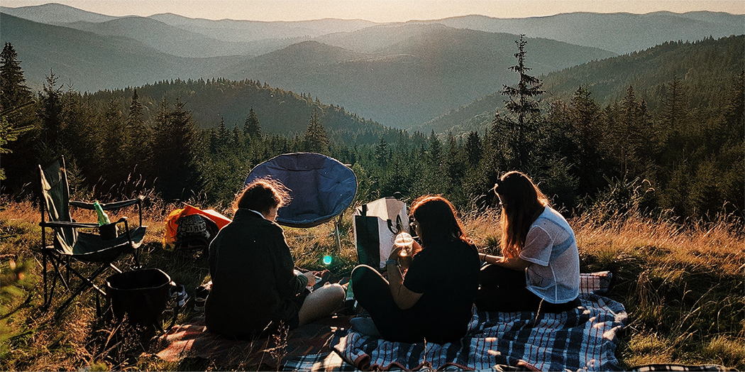 Hero image featuring three people sitting on blanket at their campsite overlooking a mountain range.