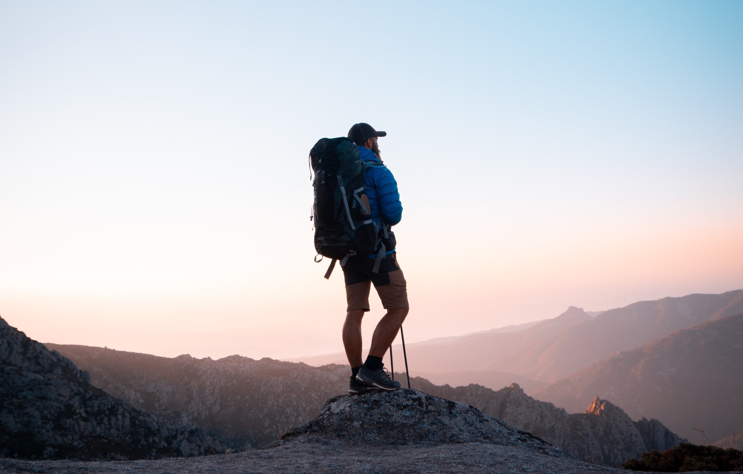 Man standing on peak with blue jacket, shorts, hiking poles and large hiking backpack