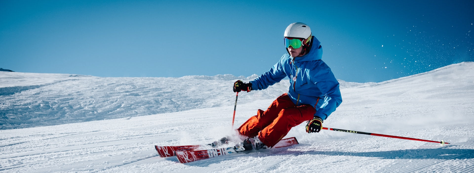 Hero image featuring a person in a blue ski jacket and red ski pants skiing down the mountain on a bluebird day.