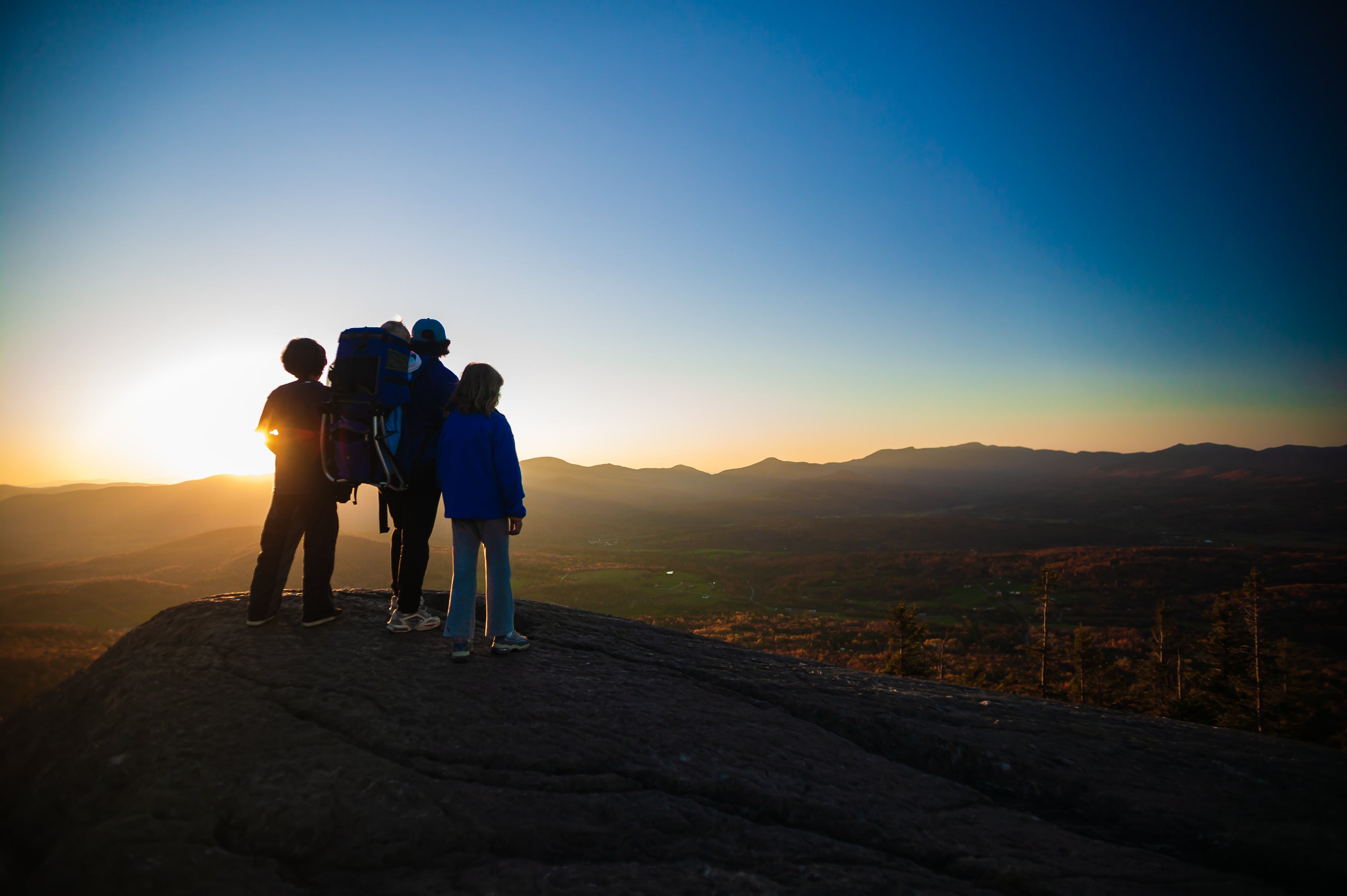 Landscape photo of a family silhouetted in hiking gear in the front left standing on a rock edge overlooking open fields, trees, and mountains way in the distance. 