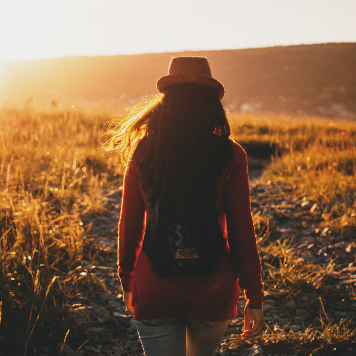 Woman walking away as the sun begins to set with a long red sweater, wearing a backpack, and hat