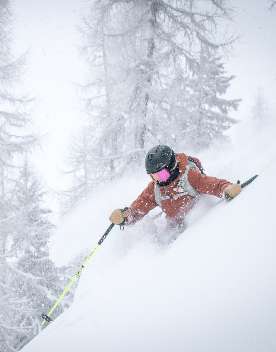 Image of a skier in deep powder doing a turn. Everything is covered in white snow but the upper half of this skier who has a brown jacket gloves, goggles, helmet and poles.
