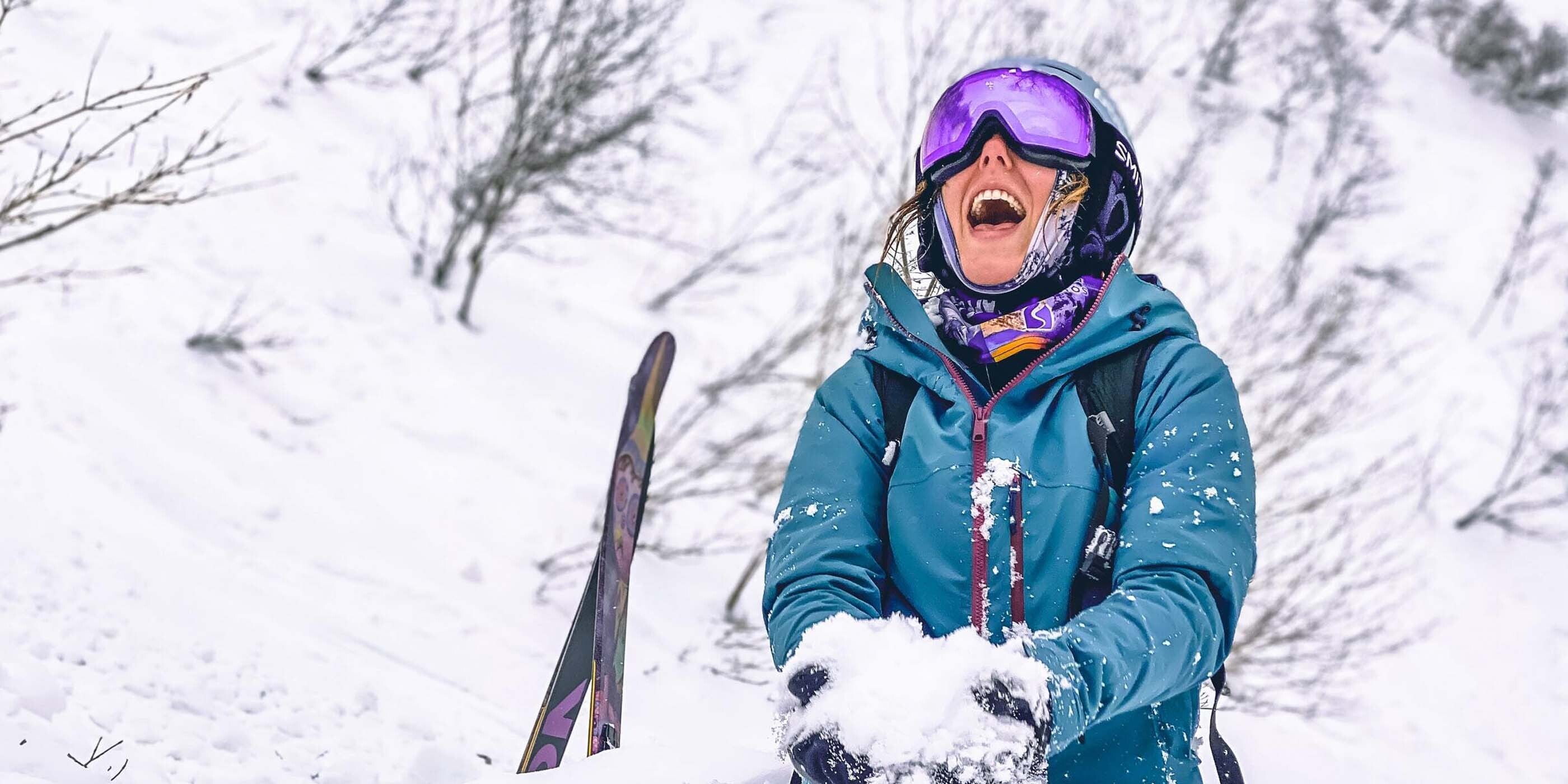 Woman in purple google and blue jacket is kneeling in snow with a huge smile holding snow in her hands, right before she's going to chuck it in the air.