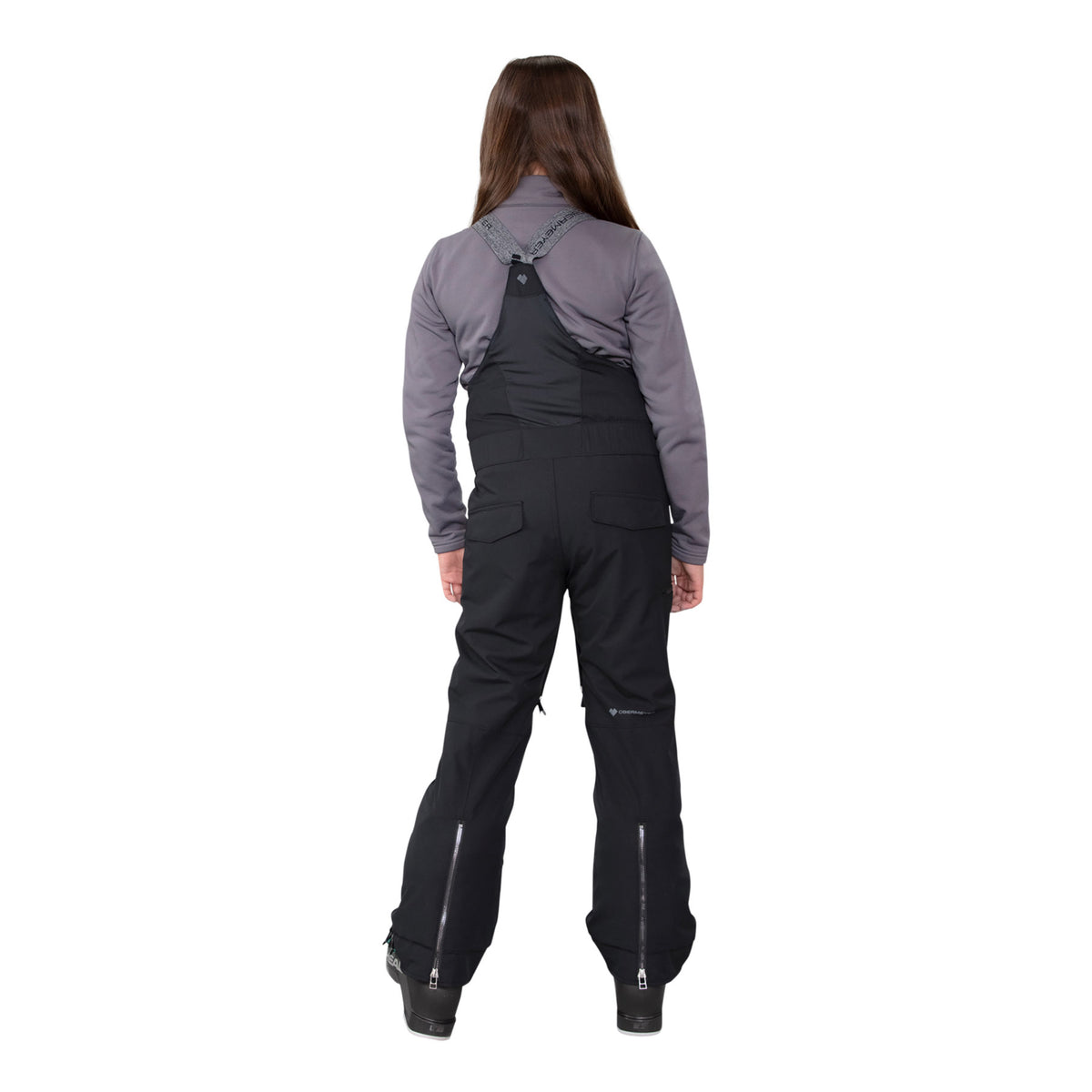Image features the back of a model wearing the Obermeyer girls Anya Bib Pant in black with black ski boots and a grey long sleeve.