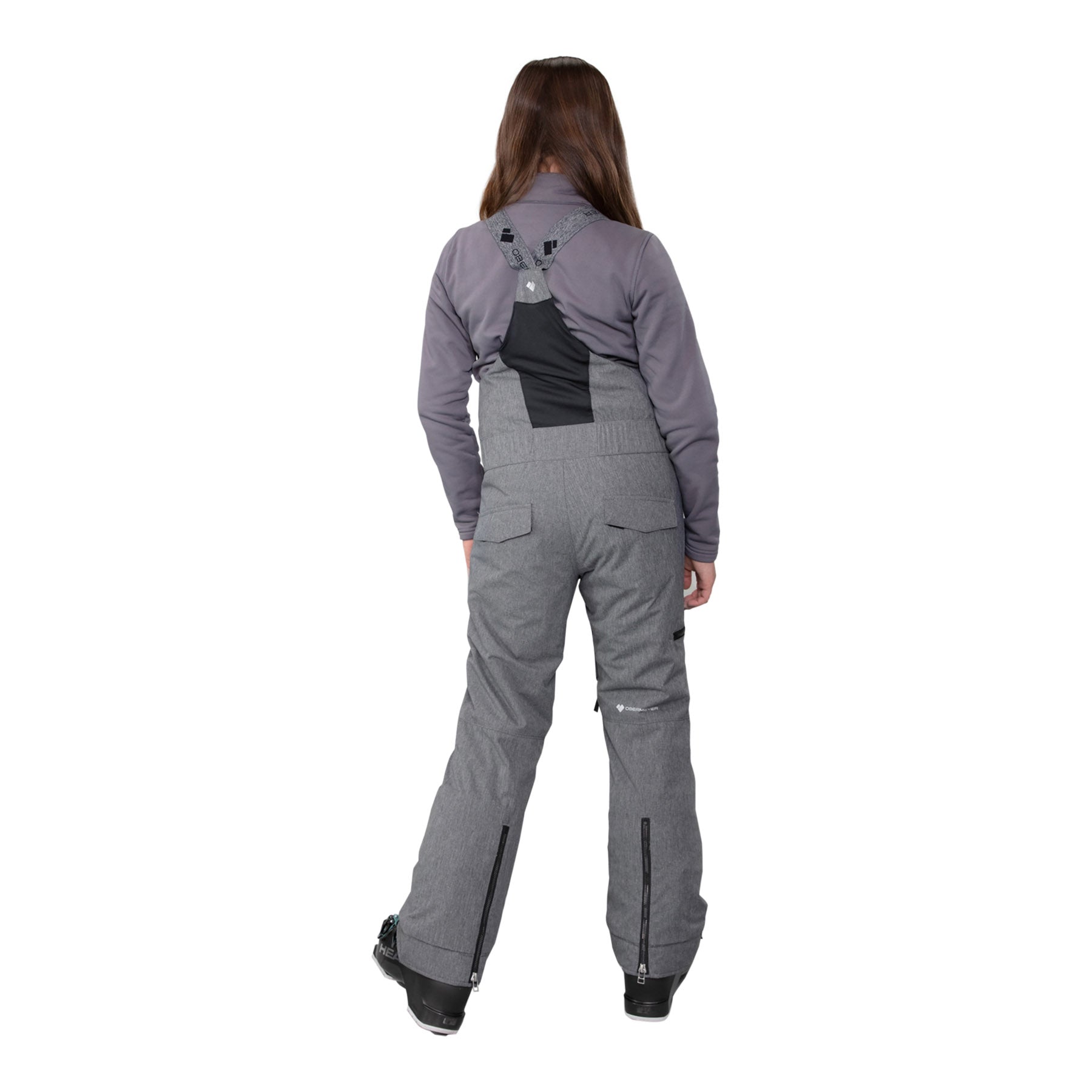 Image features a model wearing the Obermeyer girls Anya Bib Pant in light grey with black ski boots and a grey long sleeve.