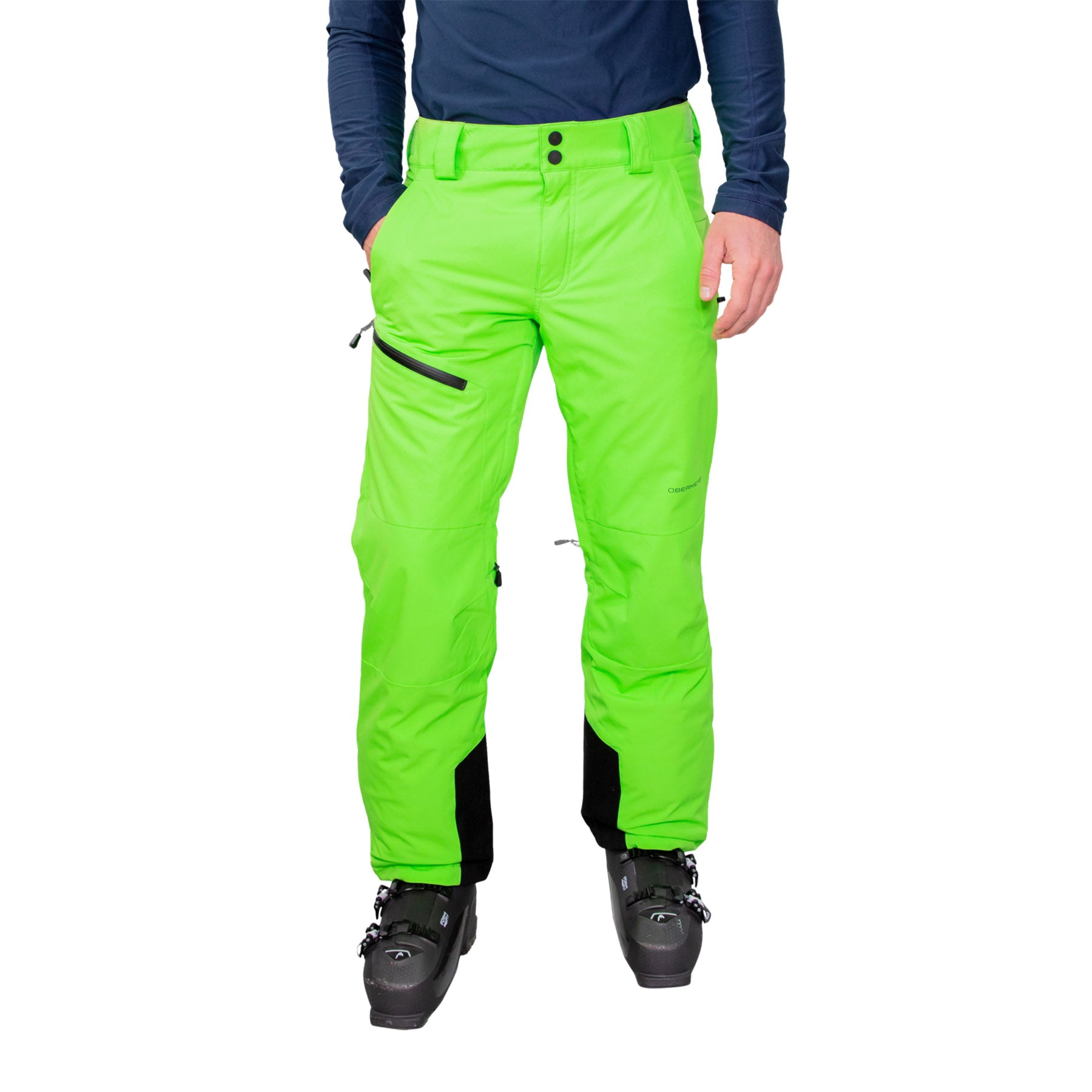 Image features a model wearing the Obermeyer Force Pant in wasabi bright green with a blue long sleeve and black ski boots.